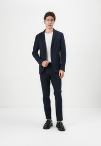 Fato azul-marinho com um corte ajustado, blazer de botão único e calças combinadas. Usado sobre uma camisa creme clara, complementado por sapatos pretos.