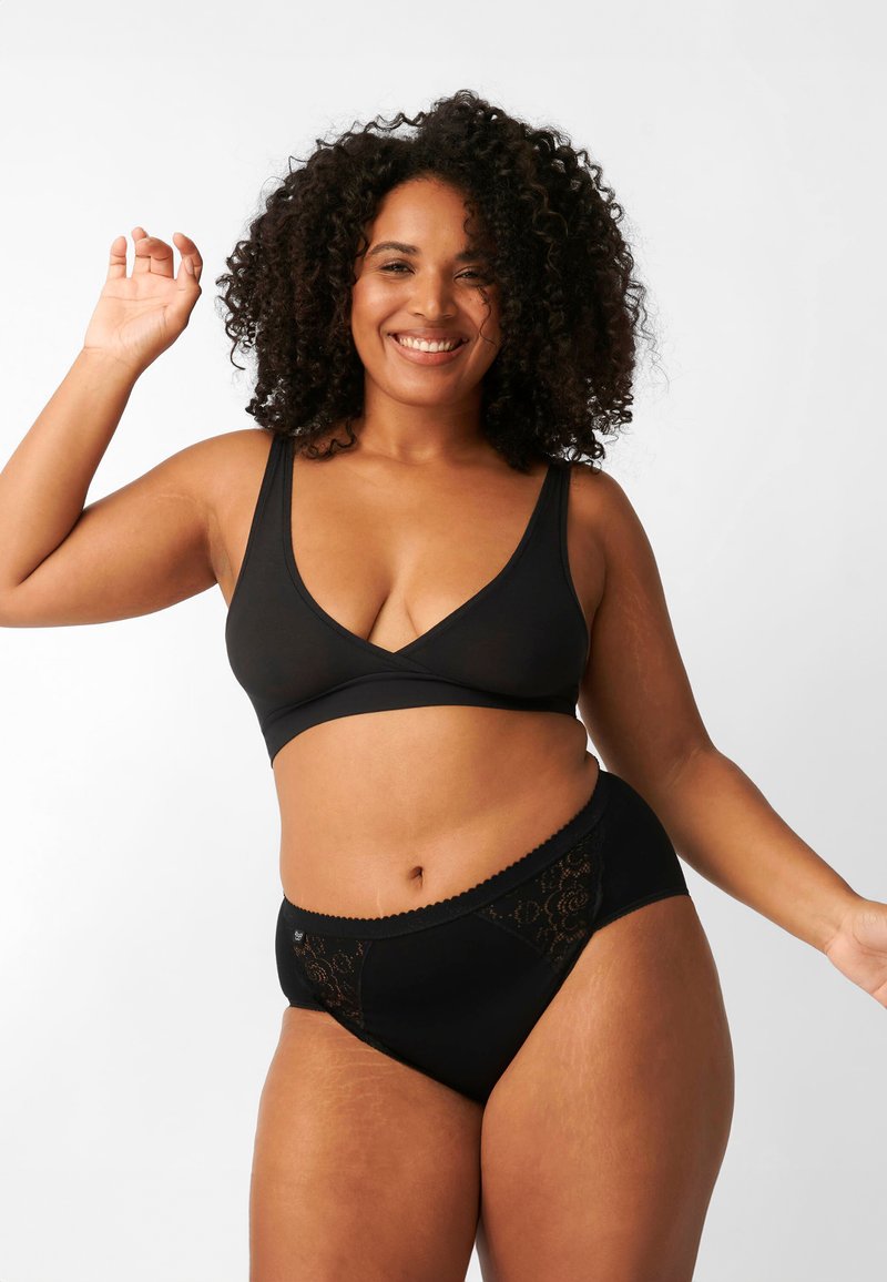 Donna sorridente con capelli ricci che indossa un reggiseno nero e biancheria intima nera con dettagli in pizzo, posando con un braccio sollevato su uno sfondo bianco.