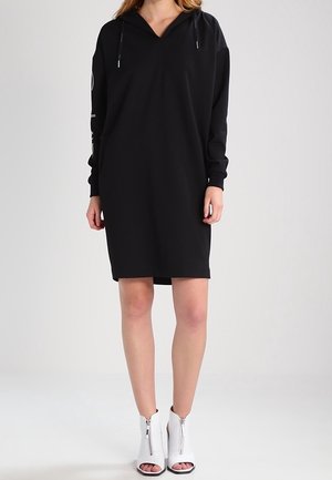 Personne portant une robe noire ample à manches longues avec capuche et cordons, associée à des bottines blanches à bout ouvert avec fermetures éclair à l'avant.