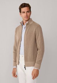Pull beige côtelé à fermeture éclair avec un col montant, porté par-dessus une chemise rayée bleu clair. Le tissu semble doux et texturé.