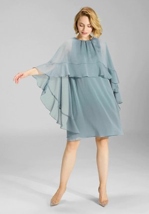 En flydende, lagdelt kjole i blødt blå stof med gennemsigtige detaljer, der har en rund halsudskæring og dramatiske kapelignende ærmer.