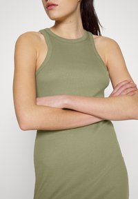 Gestreiftes, ärmelloses Kleid aus olivgrünem Stoff, mit einer figurbetonten Silhouette und rundem Ausschnitt, mit vor der Brust verschränkten Armen.