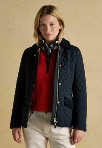 Jachetă neagră matlasată cu accesorii aurii, având guler, buzunare frontale și închidere cu fermoar, purtată peste un pulover roșu și pantaloni crem.