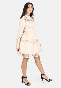 Beiges Kleid mit langen Ärmeln, hohem Kragen und aufwendigen Spitzenverzierungen. Gestufter Rüschensaum, kombiniert mit schwarzen High-Heel-Sandalen.