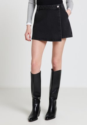 Donna che indossa una gonna a portafoglio in denim nero, stivali neri di pelle fino al ginocchio, una maglietta nera e una camicia grigia a maniche lunghe sotto.