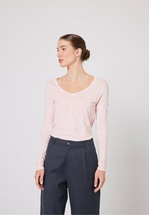 Helles rosa Langarmshirt mit V-Ausschnitt, aus einem strukturierten Stoff gefertigt. Kombiniert mit hochgeschnittenen dunkelgrauen Hosen.