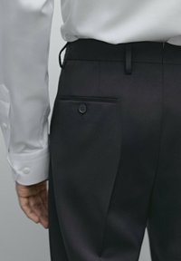 Schwarze, taillierte Hose mit einer einzelnen Gesäßtasche mit Knopf, schlankem Design und glatter Stoffqualität. Weißes Hemd teilweise sichtbar.