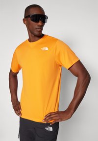 Oransje kortermet t-skjorte med rund hals og hvit logo på venstre bryst, kombinert med mørke shorts. Modellen har på seg svarte solbriller.