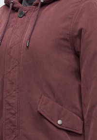 Gros plan d'une veste bordeaux avec une poche à rabat boutonnée, un bouton à l'avant et des cordons au niveau du col.