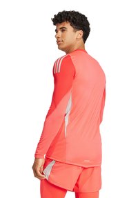 T-shirt de sport à manches longues en corail vif avec des accents gris et trois bandes sur les épaules ; tissu lisse et évacuant l'humidité.