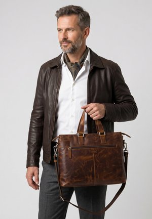 Homme portant une veste en cuir marron foncé et une chemise blanche, tenant une mallette en cuir marron avec une bandoulière, regardant sur le côté.
