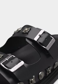 Sandalia negra con dos correas anchas que presentan hebillas plateadas y tachuelas decorativas de metal en formas florales y circulares.