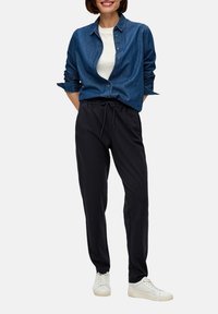 Blauwe spijkerstroven met knoopsluiting, gedragen over een wit T-shirt, gecombineerd met zwarte joggerbroeken en witte sneakers. Casual, ontspannen pasvorm.