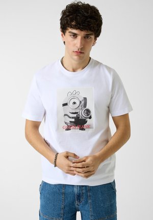 Jeune homme aux cheveux bouclés foncés portant un t-shirt blanc avec un Minion tenant un appareil photo et un jean bleu, debout devant un fond uni.