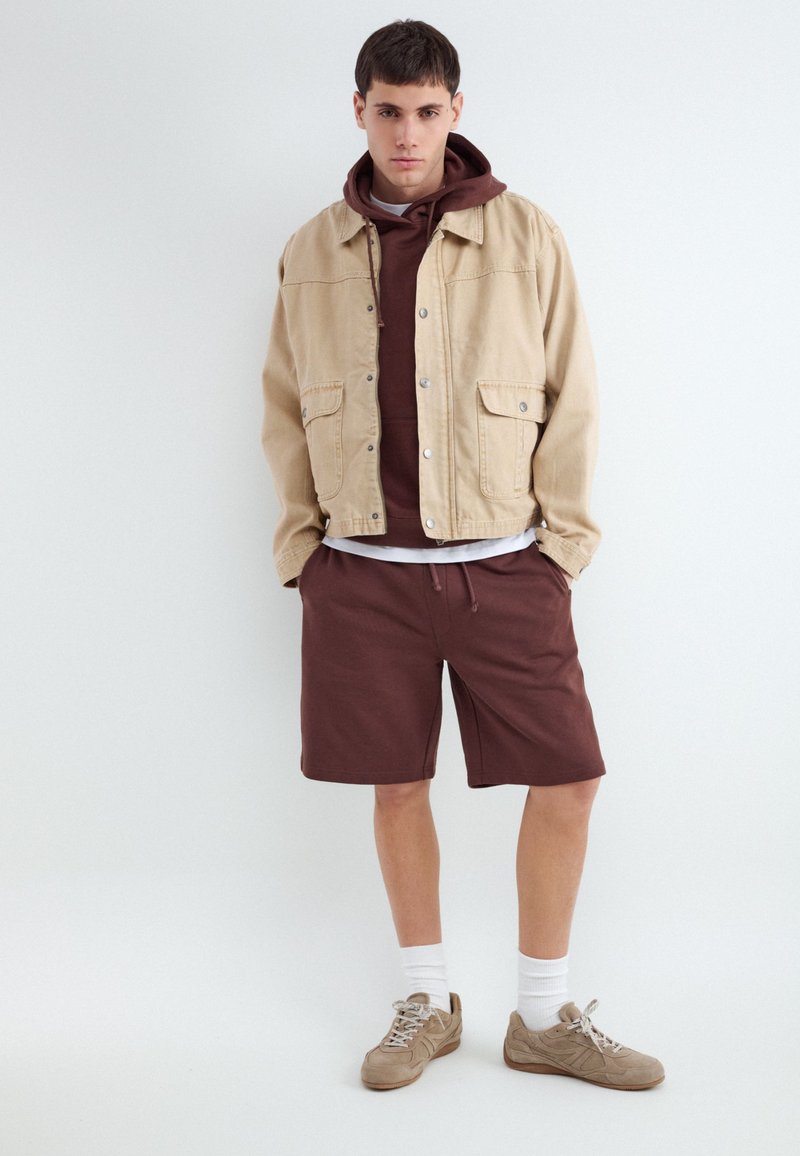Jeune homme debout portant une veste beige sur un sweat à capuche marron et un short marron assorti avec des chaussettes blanches et des baskets beiges sur fond blanc.