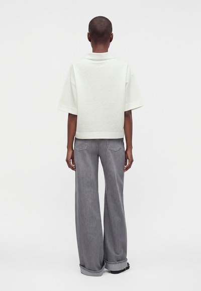 Personne de dos à la caméra, portant un haut blanc texturé à manches courtes et un jean gris large sur un fond uni.