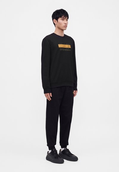 Млад мъж, облечен с черен суичър с жълто лого на Emporio Armani, черен панталон, бели чорапи и черни маратонки.