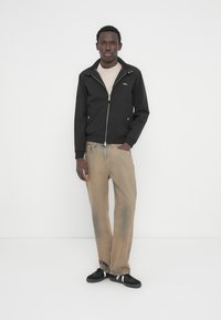 Νεαρός άνδρας που στέκεται φορώντας μαύρο μπουφάν με φερμουάρ, μπεζ μπλουζάκι, φαρδιά τζιν σε καφέ απόχρωση και μαύρα αθλητικά παπούτσια με λευκές ρίγες πάνω σε λευκό φόντο.