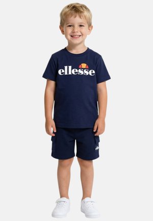Jeune garçon souriant portant un t-shirt ellesse bleu marine, un short assorti et des baskets blanches, debout devant un fond uni.