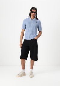Light blue knit polo with white collar, paired with black shorts and cream sneakers. Model wears dark sunglasses. Smooth texture, casual fit.