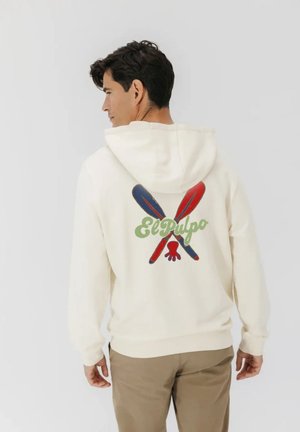 Hombre con sudadera blanca con capucha, con remos azules y rojos cruzados y texto "El Pulpo" en la espalda, de pie sobre un fondo liso.