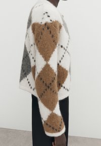 Pull en tricot avec un design court en blanc, marron et gris, orné d'un motif en losanges et de coutures noires en contour. Tissu doux et texturé.