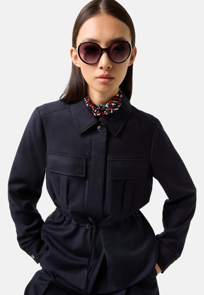 Frau mit großen runden Sonnenbrillen, einer dunkelblauen Jacke mit Fronttaschen und einem gemusterten Halstuch, die mit den Händen in den Taschen dasteht.