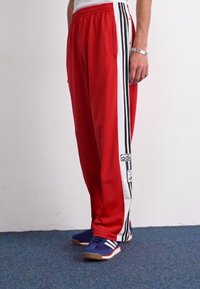 Red athletic pants with white and black stripes along the sides, featuring an elastic waistband and a logo patch on the left side.