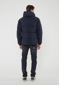 Giacca puffer blu navy con colletto alto e design trapuntato, con polsini elasticizzati e vestibilità comoda, indossata con jeans scuri e scarpe casual.