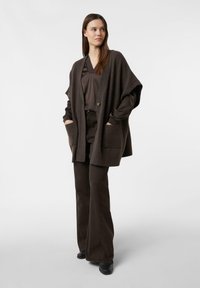 Braune Strickjacke mit übergroßer Form, tiefen Taschen und einem einzelnen Knopfverschluss, kombiniert mit dunkelbraunen, ausgestellten Hosen und schwarzen Schuhen.