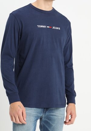 Granatowa koszula z długim rękawem z logo "TOMMY JEANS" i emblematem flagi na piersi, noszona z jasnoniebieskimi dżinsami.