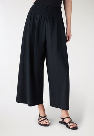 Donna che indossa pantaloni neri a gamba larga e corta con vita arricciata e sandali neri aperti con dettagli dorati.