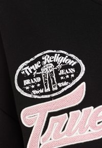 Czarny t-shirt z białym owalnym logo z napisem „True Religion” oraz grafiką dżinsów, a poniżej różowy haftowany napis.