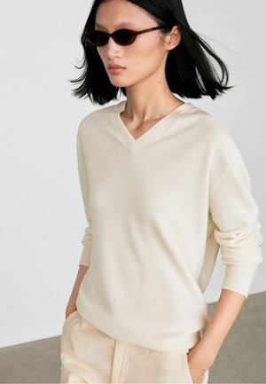 Kvinde iført små mørke solbriller og en cremefarvet V-hals sweater med hænderne i lommerne på lyse beige bukser.
