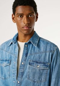 Světle modrá džínová košile s límečkem na knoflíky, dvěma náprsními kapsami a zapínáním na patentní knoflíky. Vyznačuje se vybledlou texturou a kontrastním prošíváním.