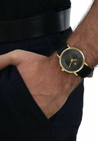 Mannelijke pols met een zwart leren horloge met gouden bezel en een donkere wijzerplaat, hand rustend in de zak van een marineblauwe broek met een zwarte riem.