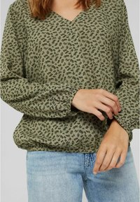 Olijfgroene blouse met een V-hals, verrijkt met een botanische print en elastische manchetten. Gestyled met casual blauwe jeans voor contrast.