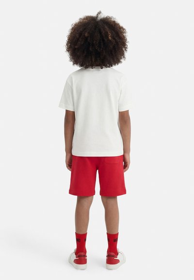 Παιδί με σγουρά μαλλιά στέκεται με την πλάτη, φορώντας λευκό μπλουζάκι, κόκκινα σορτς, κόκκινες κάλτσες και λευκά αθλητικά παπούτσια με κόκκινες λεπτομέρειες.