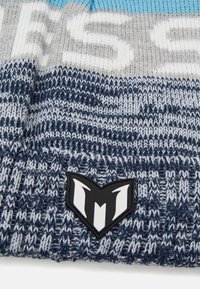Megztas skrybėlė su mėlynais, pilkais ir tamsiai mėlynais raštais, išryškintomis logotipo etiketėmis. Medžiaga yra minkšta, tekstūruota. Akcents suteikia gilumo.