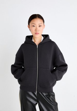 Haut à capuche noir zippé en tissu doux avec une grande capuche, deux poches avant et des poignets côtelés. Porté sur des couches grises et associé à un pantalon épuré.