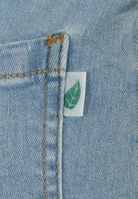 Svetlo modra denim tkanina z vidno šivano črto. Na robu je pritrjena majhna bela oznaka z zelenim logotipom lista.