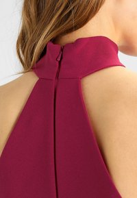 Burgunderfarbene ärmellose Bluse mit hohem Kragen und Reißverschluss auf der Rückseite. Glattes Material mit einem klaren, modernen Design und leichtem Glanz.