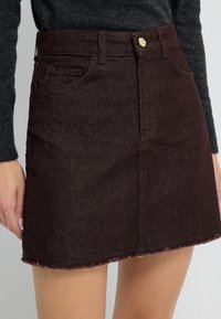 Donkerbruine denim mini-jurk met rafelige zoom, voorzakken en een gouden knoopsluiting, gedragen met een donker langemouwen top.