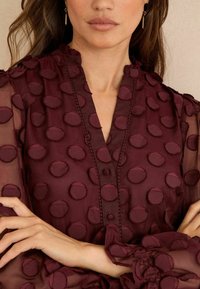 Blusa bordeaux con punti di tessuto circolari in rilievo, davanti con bottoni e maniche arricciate. Realizzata in materiale leggero e semitrasparente.