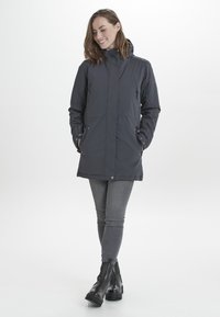Parka longue gris foncé avec une capuche, dotée de fermetures éclair et de poches. Fabriquée en matériau léger, elle a une texture lisse et une coupe décontractée.