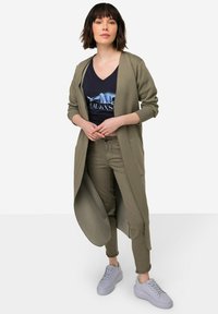 Olivově zelený dlouhý kardigan s otevřeným předním dílem, spárovaný s námořnickým tričkem s bílým grafickým designem a olivovými kalhotami, doplněný o světlé boty.