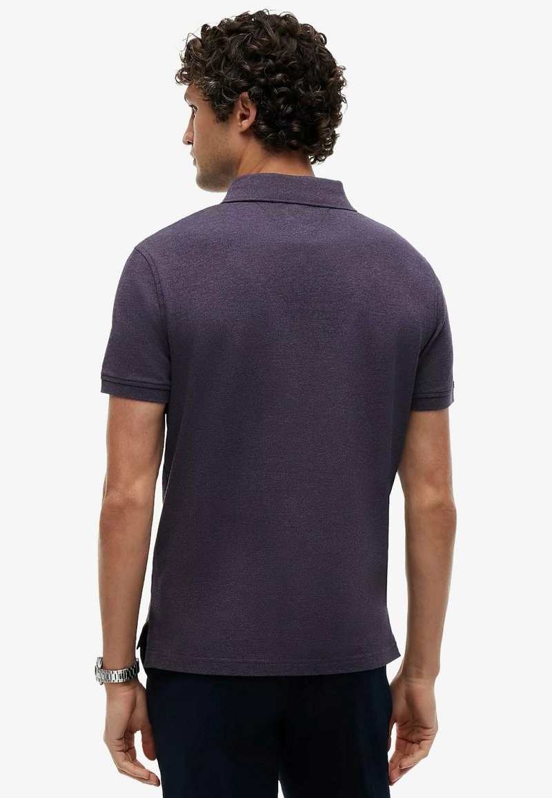 Homme aux cheveux bouclés portant un polo manches courtes violet foncé et une montre-bracelet argentée, de dos sur fond blanc.