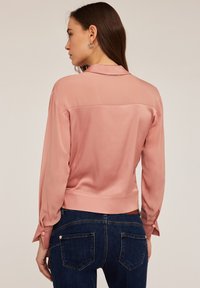 Blusa di seta rosa chiaro con maniche lunghe, polsini abbottonati e design corto, abbinata a jeans blu scuro. Trama morbida e finitura liscia.