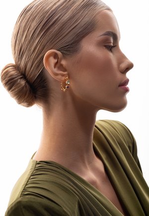 Kobieta w profilu z blond włosami związanymi w niski kok, nosząca złote kolczyki koła z kolorowymi kamieniami i oliwkowo zielony marszczony top.