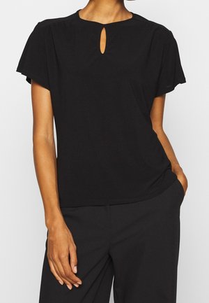 Kvinde iført en sort top med korte ærmer og en lille dråbeformet udskæring nær halsudskæringen, kombineret med sorte bukser, hånd i lommen.